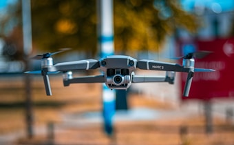 A drone, specifically a Mavic 2, is captured mid-flight with a blurred background of trees and a bright blue sky. The focus on the drone highlights its propellers and camera, suggesting a stable hover. The setting appears sunny, accentuating the metallic surface of the drone.
