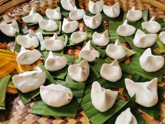 A collection of white, steamed dumplings placed on pieces of green banana leaves, arranged on a wicker basket background. The dumplings have a folded, crescent shape and appear freshly prepared.