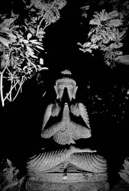 A stone statue of Buddha is seated in a meditative posture with hands in a prayer position. The figure is surrounded by lush foliage, presenting an intricate play of light and shadow, which highlights the serene expression of the sculpture.