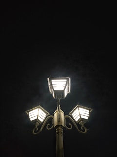 Three illuminated streetlamp lights are positioned on a single pole against a dark night sky. The lamps have a vintage design with decorative elements.