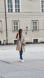 A woman in a light beige trench coat walking through a softly lit urban street with neutral-toned buildings.