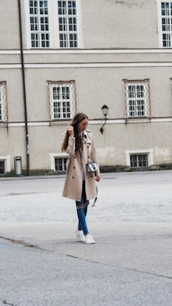 A woman in a light beige trench coat walking through a softly lit urban street with neutral-toned buildings.