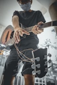 A person wearing a face mask and glasses is holding a guitar with strings hanging loosely. The perspective is from below, emphasizing the guitar and their outstretched arm. The background is blurred, giving focus to the individual and the guitar.