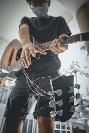 A person wearing a face mask and glasses is holding a guitar with strings hanging loosely. The perspective is from below, emphasizing the guitar and their outstretched arm. The background is blurred, giving focus to the individual and the guitar.