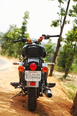 A motorcycle is parked on a sandy path with trees in the background. The license plate bears text and a sketch, and there are marigold garlands hanging from the handlebars.