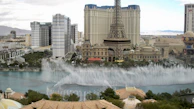 A cityscape featuring a tall structure resembling the Eiffel Tower surrounded by ornate buildings and modern high-rise hotels. In the foreground, fountains spray water high into the air in an elaborate display over a large water body. The setting appears to be an urban tourist destination with a mix of architectural styles.
