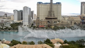 A cityscape featuring a tall structure resembling the Eiffel Tower surrounded by ornate buildings and modern high-rise hotels. In the foreground, fountains spray water high into the air in an elaborate display over a large water body. The setting appears to be an urban tourist destination with a mix of architectural styles.