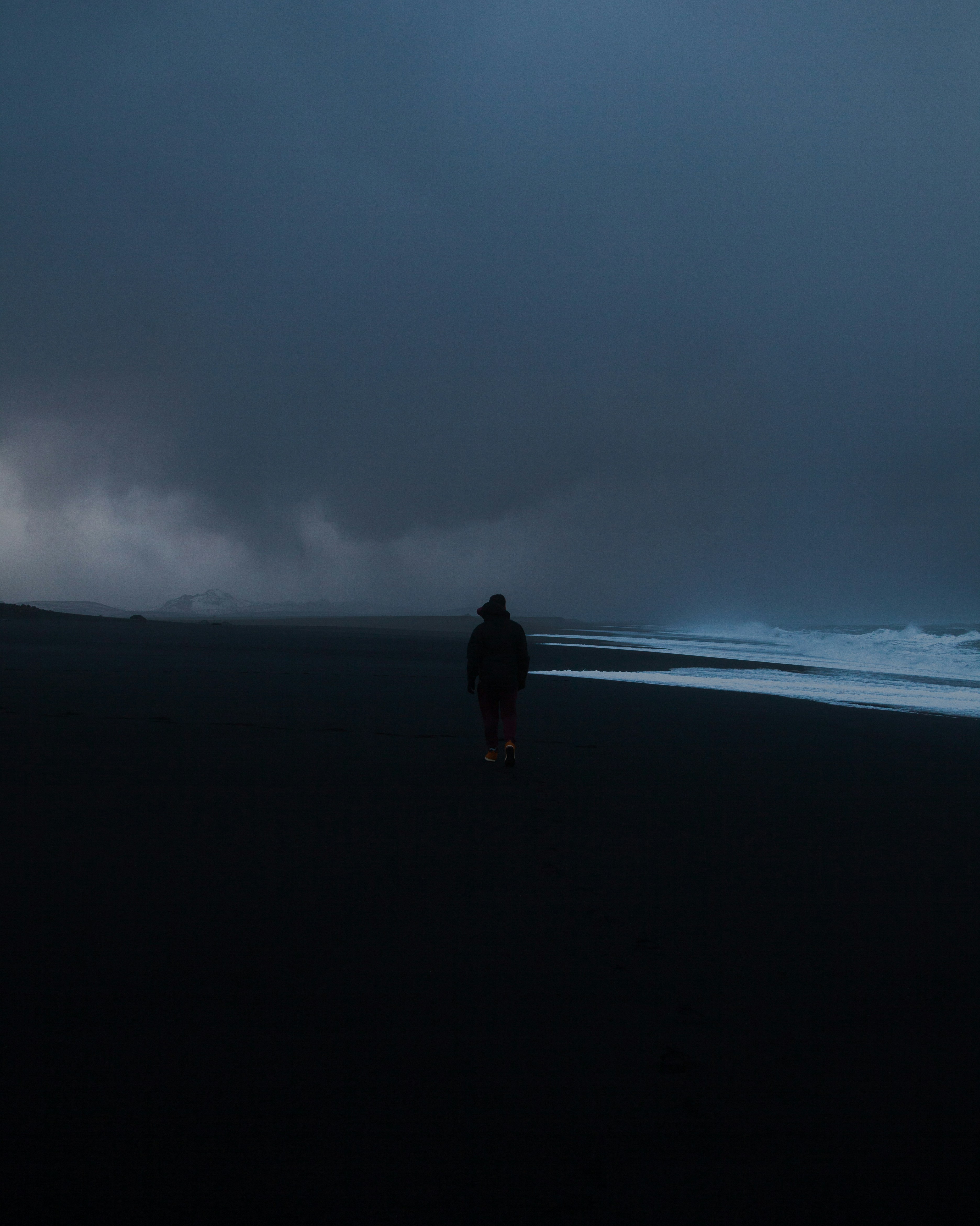man in black jacket walking on black sand under gray cloudy sky