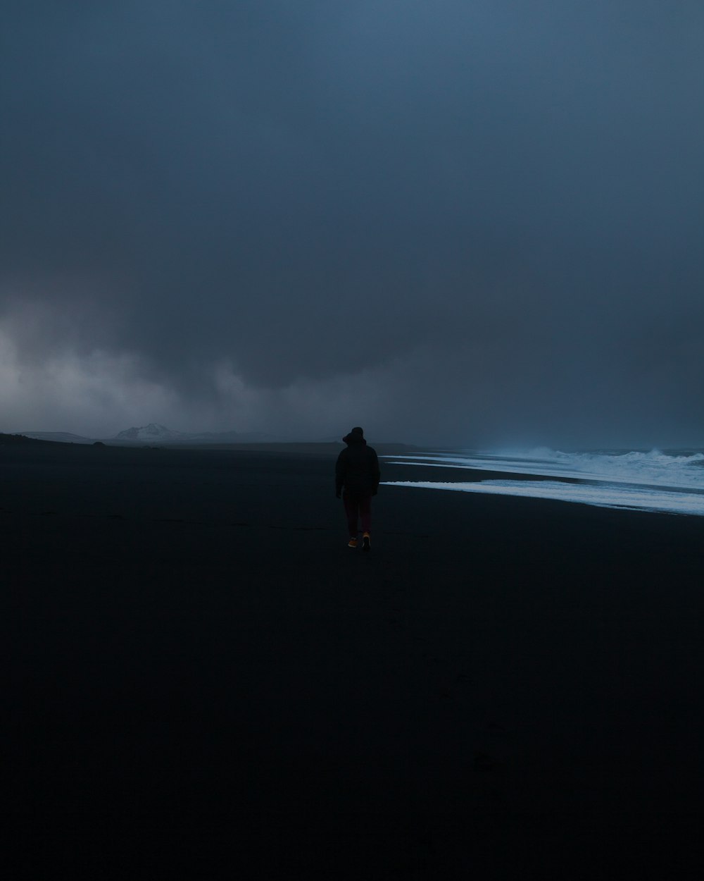 man in black jacket walking on black sand under gray cloudy sky