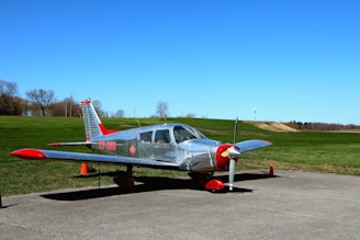 A small silver aircraft with red accents is parked on a concrete surface. The plane is positioned in front of a wide expanse of manicured green grass and a clear, vibrant blue sky. There are orange traffic cones around the aircraft and leafless trees in the background, suggesting an autumn or early spring setting.