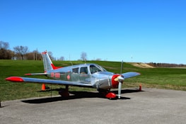 A small silver aircraft with red accents is parked on a concrete surface. The plane is positioned in front of a wide expanse of manicured green grass and a clear, vibrant blue sky. There are orange traffic cones around the aircraft and leafless trees in the background, suggesting an autumn or early spring setting.