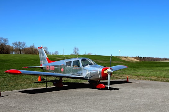 A small silver aircraft with red accents is parked on a concrete surface. The plane is positioned in front of a wide expanse of manicured green grass and a clear, vibrant blue sky. There are orange traffic cones around the aircraft and leafless trees in the background, suggesting an autumn or early spring setting.