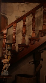 A wooden staircase with intricately carved balusters and a decorative sculpture of a cherub holding a cup. The stone wall background accentuates the classical and rustic aesthetic.