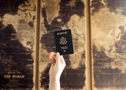 A hand is holding a United States passport in front of a blurred background showing a vintage-style world map with sepia tones.