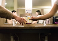 Two individuals are in a restroom, with one person handing a sanitary pad to the other. They are standing near a sink with mirrors and a hand sanitizer dispenser is visible in the background.