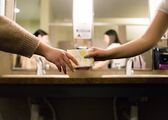 Two individuals are in a restroom, with one person handing a sanitary pad to the other. They are standing near a sink with mirrors and a hand sanitizer dispenser is visible in the background.
