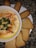 An appetizing bowl of hummus garnished with chickpeas, paprika, and fresh parsley is placed on a white plate. Surrounding the bowl are pieces of toasted bread and crispy breadsticks, all set against a dark granite surface.