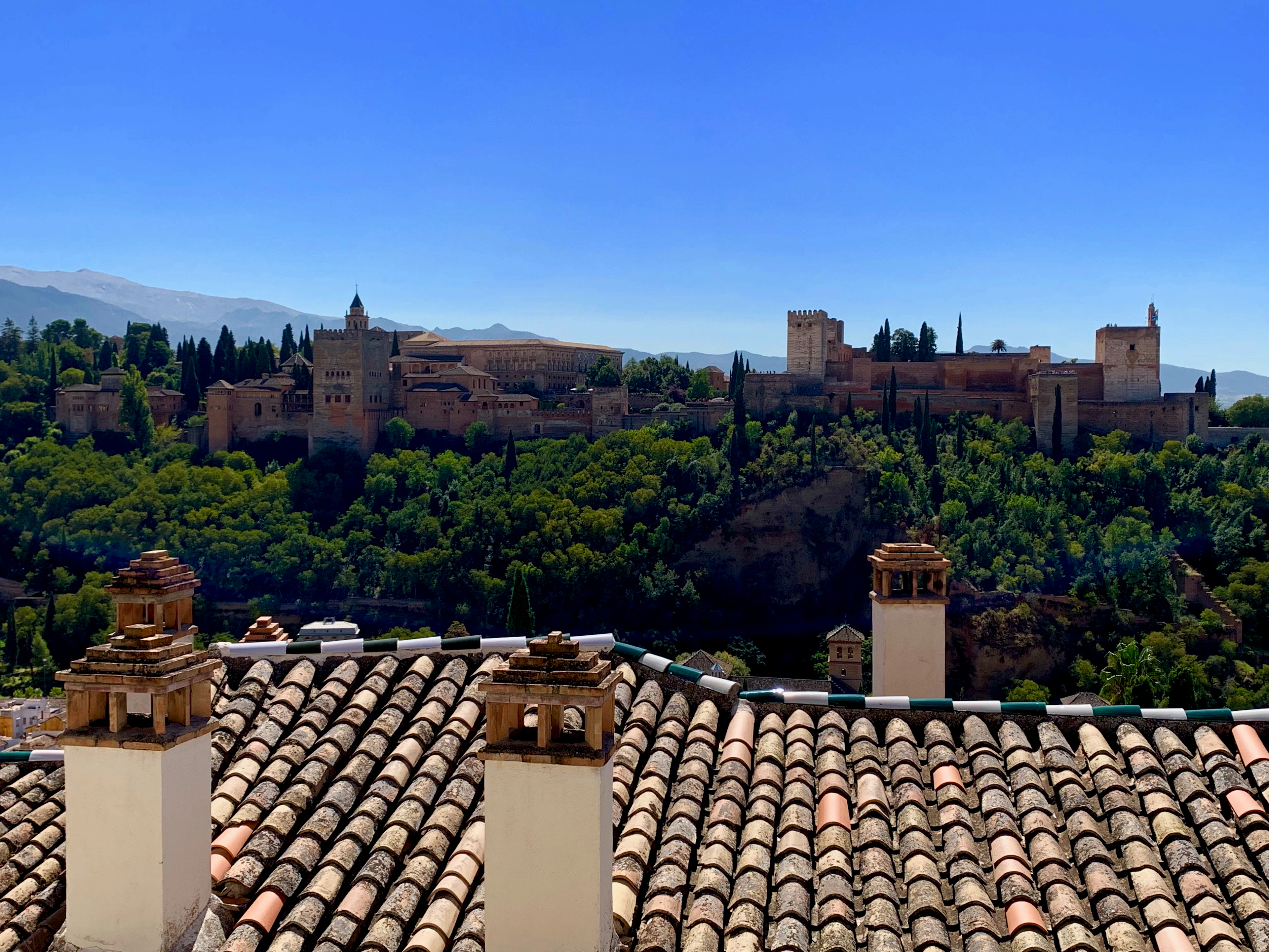 Traditional clay chimney pots frame a rooftop view of the Alhambra fortress against a clear blue sky.