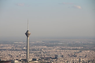 A tall communication tower dominates the skyline of a sprawling cityscape, with densely packed buildings extending into the distance. The sky appears clear with a few scattered clouds.