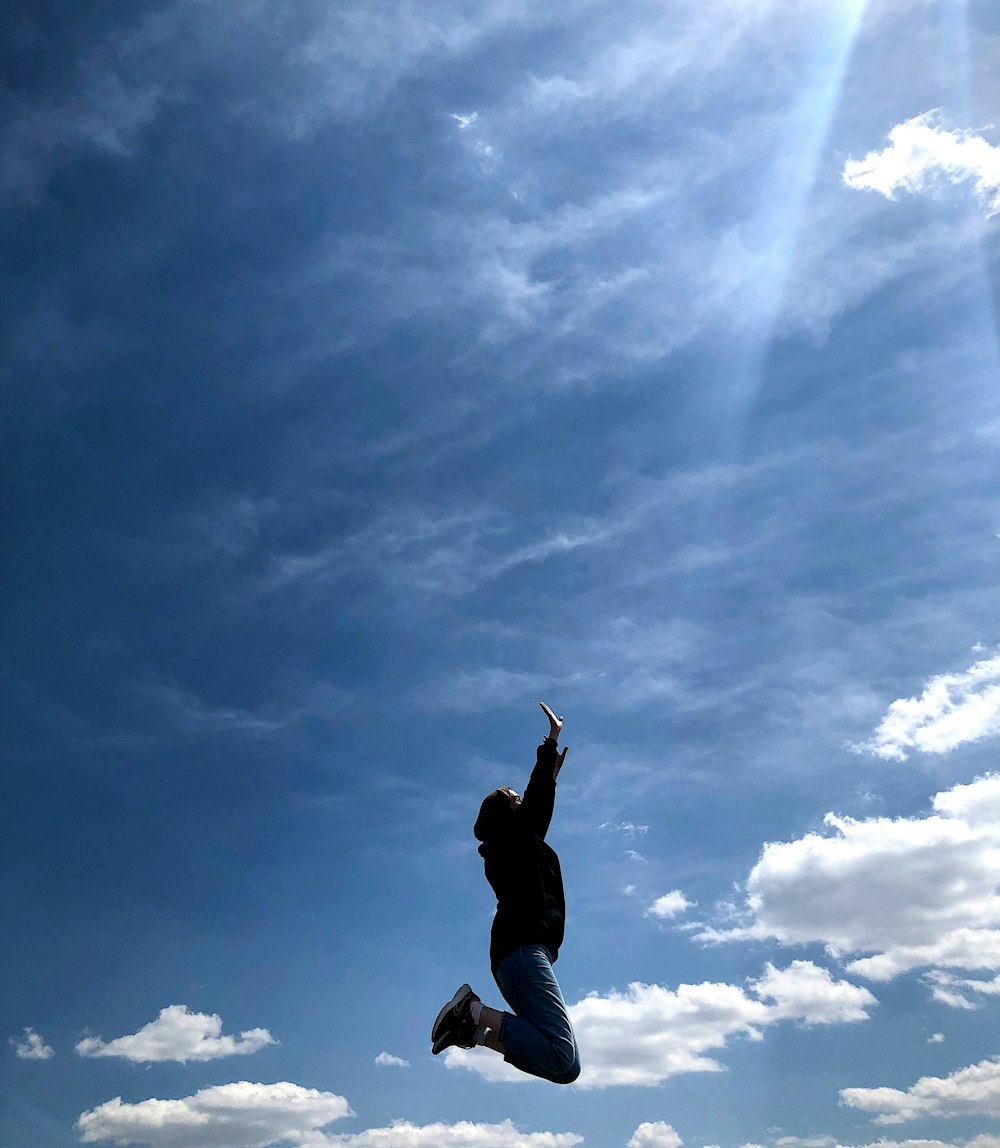 man in black jacket and blue denim jeans jumping under blue and white sunny cloudy sky