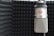 A professional microphone is positioned in an acoustic foam-lined room, likely a recording studio. The microphone has a metallic mesh grille and a red rectangular logo. The surrounding walls are covered in dark acoustic foam panels with a pyramid pattern to manage sound reflections.