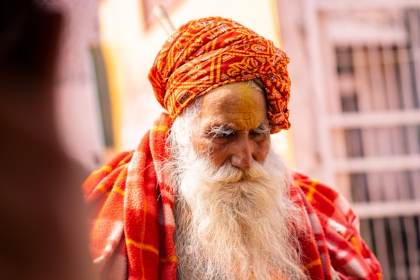 An elderly man with a long white beard wears a vibrant orange-red patterned turban. His face is partially painted with bright yellow pigments, and he is cloaked in a colorful red and orange fabric. The blurred background suggests an outdoor setting.