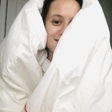 A person is wrapped warmly in a white comforter, with only their face partially visible. The expression appears relaxed and cozy.