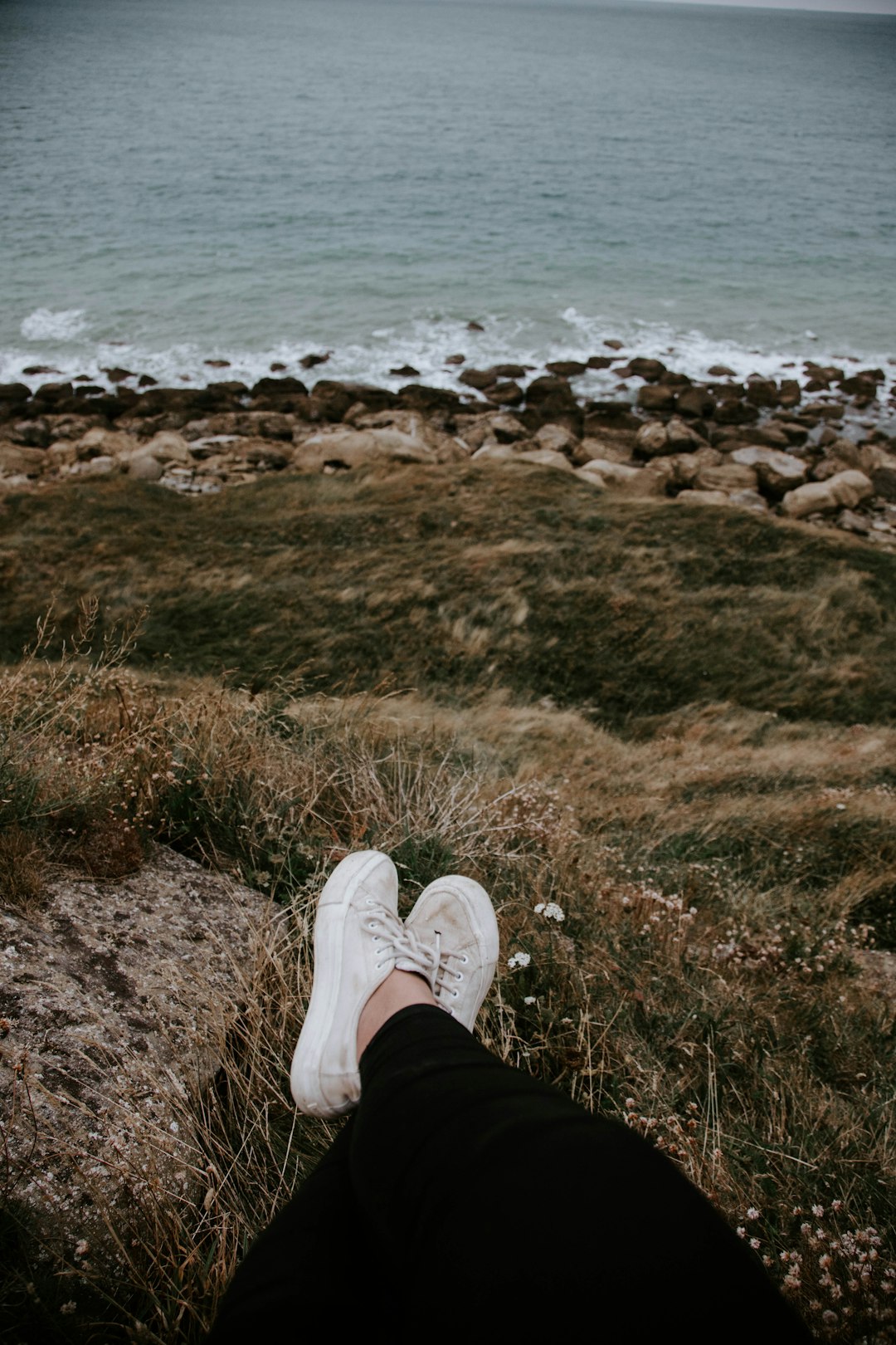 person in black pants and white sneakers sitting on brown grass near body of water during