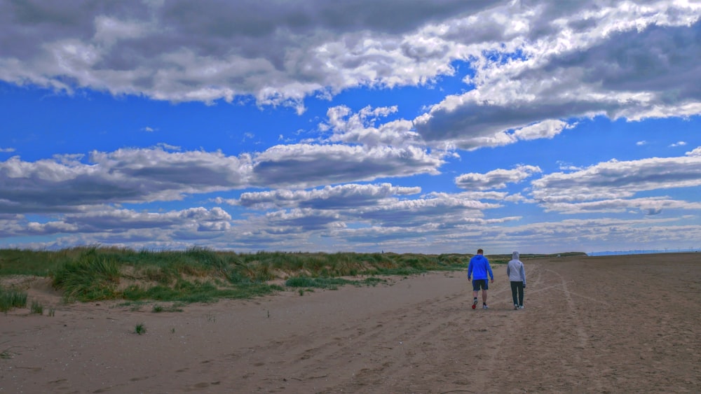2 Männer spazieren tagsüber auf braunem Sand unter blauem Himmel und weißen Wolken