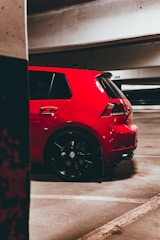 A vibrant red hatchback car is parked in an underground parking garage. The car has sporty features with black rims and visible branding on the wheels. The setting is dimly lit, with concrete walls and parking lines visible on the asphalt floor.