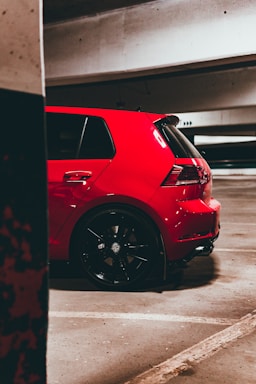 A vibrant red hatchback car is parked in an underground parking garage. The car has sporty features with black rims and visible branding on the wheels. The setting is dimly lit, with concrete walls and parking lines visible on the asphalt floor.