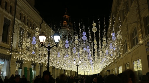 A street at night is adorned with numerous hanging decorative lights, creating a festive atmosphere. The lights are arranged in cascading patterns, with some larger spherical lights interspersed among smaller lights. Street lamps and the silhouettes of people walking are visible. The buildings lining the street are illuminated by the festive lights.