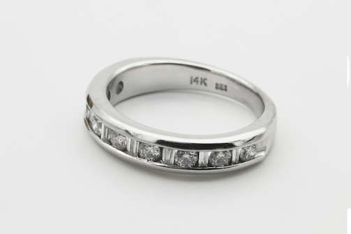 A silver ring with a row of seven small, sparkling diamonds embedded along its band. The interior of the band is inscribed with '14K'. The ring is simple yet elegant, showcasing a minimalist design.
