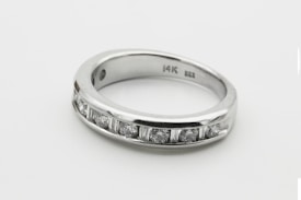 A silver ring with a row of seven small, sparkling diamonds embedded along its band. The interior of the band is inscribed with '14K'. The ring is simple yet elegant, showcasing a minimalist design.