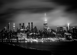 A monochrome cityscape of New York City at night with the skyline prominently featuring tall skyscrapers, including the illuminated Empire State Building. The city lights reflect softly on the water in the foreground, creating a serene and majestic urban scene.