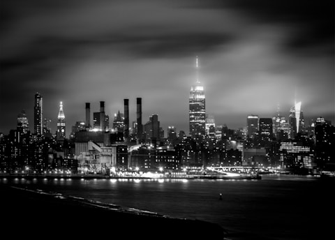 A monochrome cityscape of New York City at night with the skyline prominently featuring tall skyscrapers, including the illuminated Empire State Building. The city lights reflect softly on the water in the foreground, creating a serene and majestic urban scene.