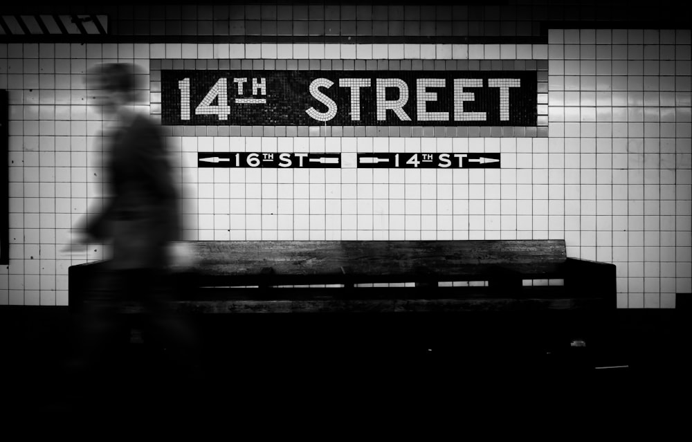 a black and white photo of a man walking past a subway sign