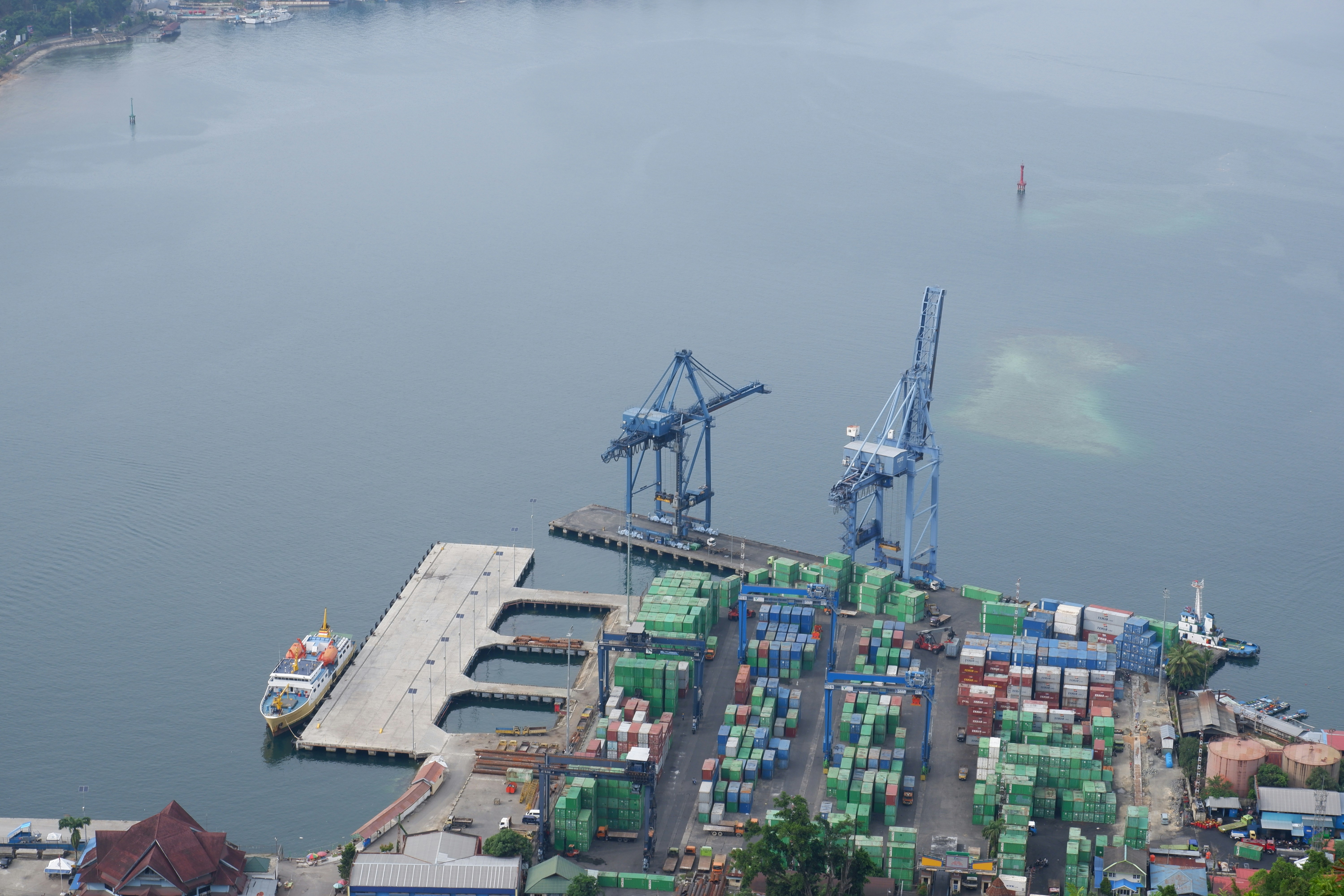 Stacked shipping containers at a busy port terminal, muted grey and blue tones, industrial overhead cranes visible