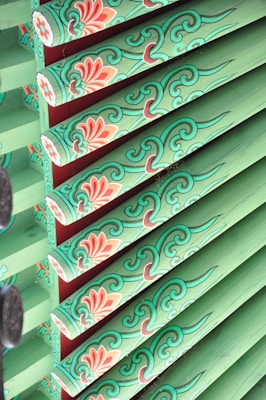 Intricate wooden beams decorated with traditional floral patterns are painted in vibrant green and accented with red and white floral motifs. The designs feature swirling lines and detailed blossoms, highlighting craftsmanship.