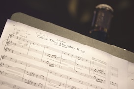 A close-up view of a sheet music titled 'Come Thou Almighty King' laid out on a music stand, with a microphone blurred in the background. Handwritten notes and annotations are visible on the sheet music.