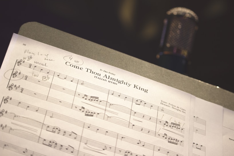 A close-up view of a sheet music titled 'Come Thou Almighty King' laid out on a music stand, with a microphone blurred in the background. Handwritten notes and annotations are visible on the sheet music.
