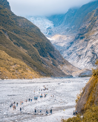 A scenic mountainous landscape with a glacier visible in the background. A large group of people is walking across a rocky terrain in the valley, surrounded by steep hills covered in vegetation. The sky appears overcast with low-hanging clouds.
