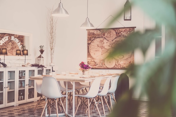 A dining area featuring a modern wooden table surrounded by white chairs. A large, vintage-style world map serves as a wall decoration. Soft lighting is provided by pendant lamps, and decorative items such as plants and a flower arrangement on the table enhance the ambiance. A sideboard displays dishware and is adorned with more decorative elements.