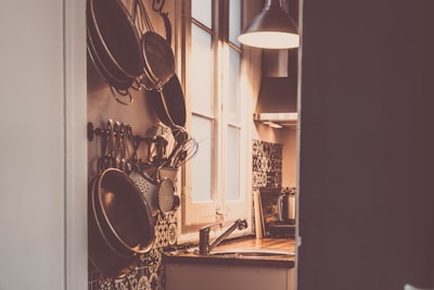 A cozy kitchen environment features a window letting in warm light, illuminating hanging utensils and pans on the wall. The kitchen counter is wooden, and beneath a hanging lamp, there is a metal sink. The atmosphere has a rustic feel with patterned tiles adding to the aesthetic.