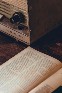 A vintage radio with a visible dial sits next to an open book, both resting on a wooden surface. The radio has an old-fashioned design with the label 'All Transistor' shown at the bottom. The book is open to a page filled with text, suggesting an atmosphere of nostalgia or history.