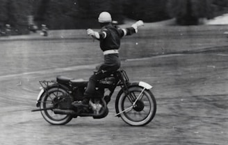 A person riding a vintage motorcycle with arms outstretched, possibly performing a balancing act or stunt. The image is in black and white, adding a classic and nostalgic feel. The motorcycle appears to be moving across a textured surface with blurred trees in the background.