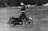 A person riding a vintage motorcycle with arms outstretched, possibly performing a balancing act or stunt. The image is in black and white, adding a classic and nostalgic feel. The motorcycle appears to be moving across a textured surface with blurred trees in the background.