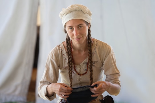 A person with long braided hair and a headscarf is wearing traditional-style clothing with decorative stitching. They are holding a piece of fabric or clothing. The background is a light-colored fabric, possibly a tent.