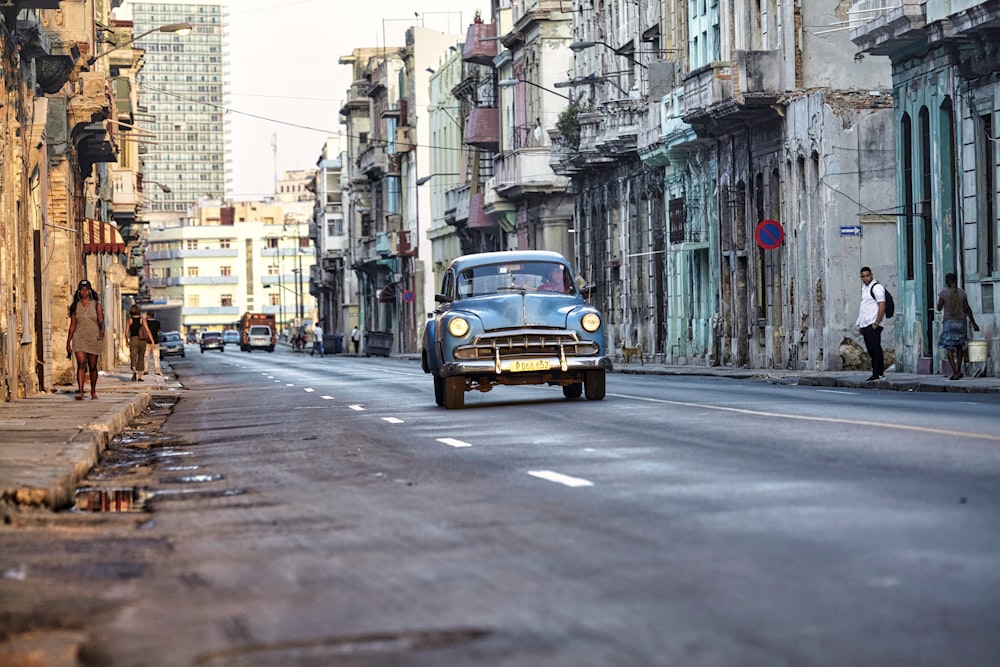 an old blue car driving down a street next to tall buildings