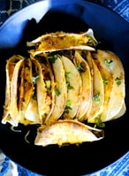 A collection of tacos arranged on a dark plate. The tacos are filled with melted cheese, vegetables, and garnished with small greens. The vibrant ingredients appear fresh and appetizing.